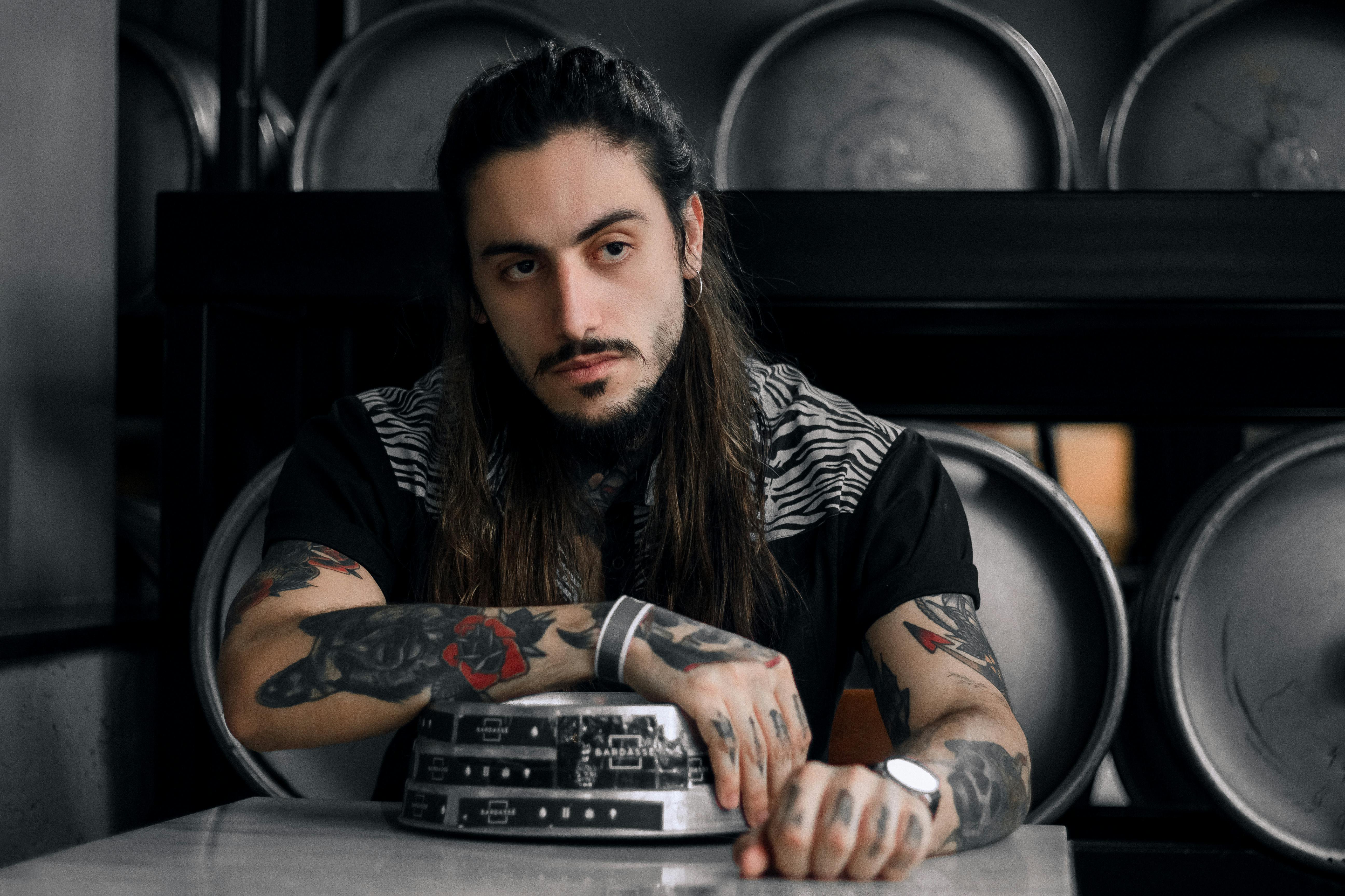 Man With Long Hair And Tattoos Sitting On A Chair Behind A Table · Free Stock Photo
